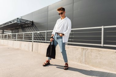 Siyah bir bez çanta ile trendy sandalet şık yaz giysileri Avrupa Genç adam hipster güneşli bir gün açık havada pozlar. Şehirde güneş gözlüğü çekici adam. Yaz moda erkek giyim.