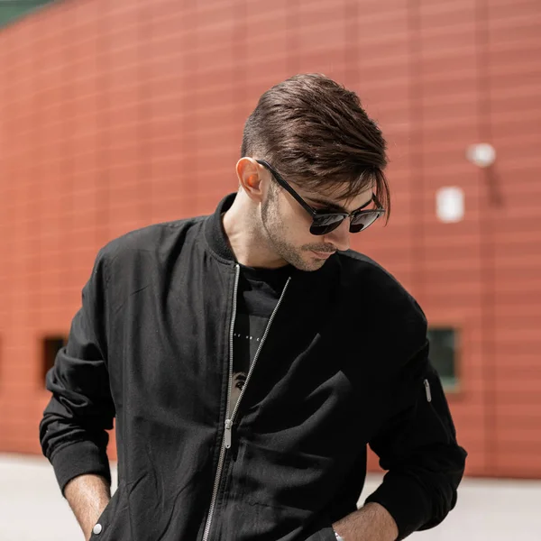 Trendy bir saç stili ile şık bir siyah ceket moda güneş gözlüğü Amerikan Genç hipster adam bir yaz gününde kırmızı modern bina yakın rahatlatır. Yakışıklı adam yürüyüş hoşlanır.