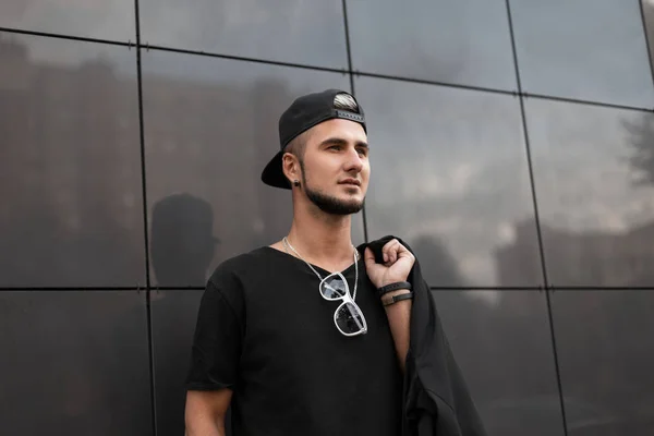 Ein gutaussehender junger Mann mit trendiger Baseballkappe, schwarzem T-Shirt, Sonnenbrille und Bart steht an einer modernen grauen Wand auf der Straße. amerikanischer stylischer Hipster in der Stadt. — Stockfoto