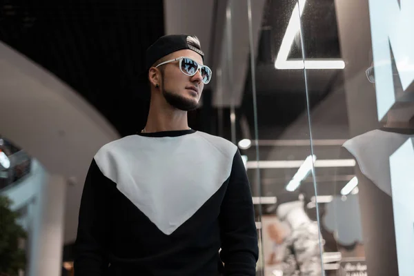 Cool Young hipster man in een modieuze zwarte pet in een stijlvolle Sweatshirt in Vintage witte zonnebrillen staat in een moderne winkel. Aantrekkelijke Urban Guy met een baard binnenshuis. Trendy herenkleding. — Stockfoto