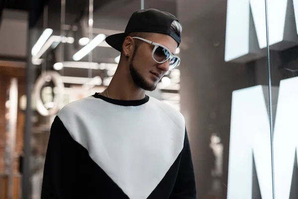Jonge modieuze hipster man in stijlvolle zonnebrillen in een trendy Sweatshirt in een Amerikaanse Baseball Cap staat in een winkel in de buurt van een helderglazen vitrine. Knappe moderne kerel lopen rond de winkel. — Stockfoto