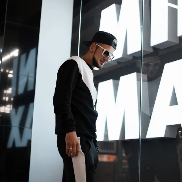 Νέος ελκυστικός άντρας με γένια με μούσι σε μαύρο καπέλο εκλεκτής ποιότητας σε κομψά γυαλιά ηλίου σε ένα μαύρο μοδάτο Αθλητικό κοστούμι περπατάει μέσα από ένα εμπορικό κέντρο. Ωραίος τύπος στο εσωτερικό. Μοντέρνα ανδρική — Φωτογραφία Αρχείου