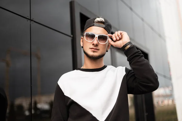 El joven y moderno hombre hipster con barba en una elegante gorra negra con una sudadera blanco-negra se levanta y endereza las gafas de sol cerca de una pared gris al aire libre. Modelo de tipo urbano descansando en la ciudad. Estilo de calle . — Foto de Stock