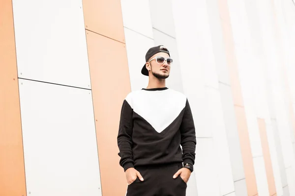 Jonge knappe hipster man in een modieuze zwarte pet in een stijlvolle Sweatshirt in broek in witte zonnebrillen staat in de buurt van een modern gebouw op een heldere zomerdag. Aantrekkelijke Urban Guy met een baard. — Stockfoto