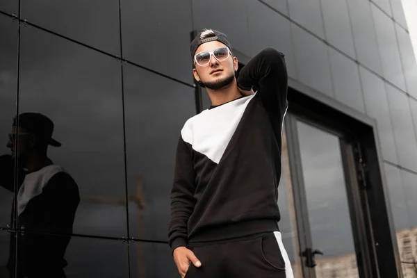 Urban Young hipster man in stijlvolle zwarte pet in trendy zonnebrillen in modieuze Sweatshirt poses in de buurt van een grijze muur in de stad. Knappe kerel met een stad op een straat in de zomerdag. Streetstyle. — Stockfoto