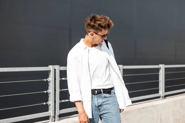 Městský mladý hipíšský muž v elegantní bílé košili v tričku ve starožitných džínách v moderních slunečních brýlích prochází městem poblíž šedé budovy. Moderní člověk na procházce za slunečného dne. — Stock fotografie