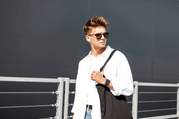 Joven con estilo en camisa blanca de moda en gafas de sol de moda en vaqueros azules vintage con una bolsa de tela negra posando en un día soleado cerca de un edificio gris al aire libre. Chico moderno en la calle. Estilo juvenil — Foto de Stock