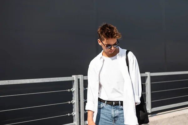 Young model man with hairstyle in stylish white shirt in t-shirt in blue jeans in trendy sunglasses with a cloth bag walk near a gray building on sunny day. Hipster boy. Fashionable menswear.