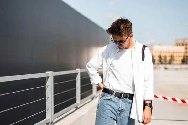 Młody modny model człowiek w T-shirt w białej koszulce w stylu Vintage niebieskie dżinsy w okulary spacerowe wokół miasta w pobliżu szarego budynku. Europejski facet Hipster cieszy słoneczny dzień. Młodzieżowy styl uliczny. — Zdjęcie stockowe