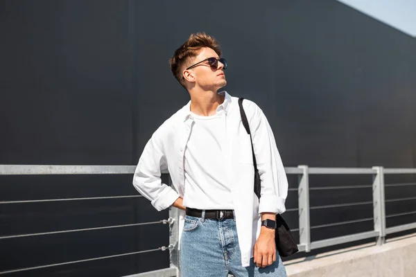 Elegante jovem hipster em óculos escuros em roupas brancas da moda com penteado moderno com saco posando perto de um edifício moderno em um dia ensolarado ao ar livre. Um modelo urbano. Estilo de juventude de verão . — Fotografia de Stock