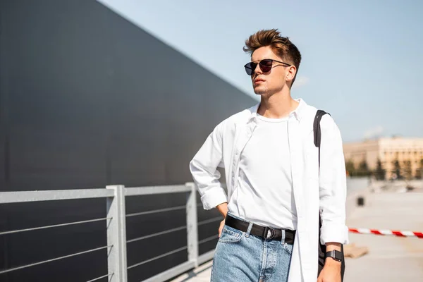 Urbano jovem hipster homem em roupas brancas elegantes em jeans na moda em óculos de sol vintage com uma bolsa vintage preta relaxa perto de edifício moderno na cidade. Homem americano na rua em um dia ensolarado . — Fotografia de Stock