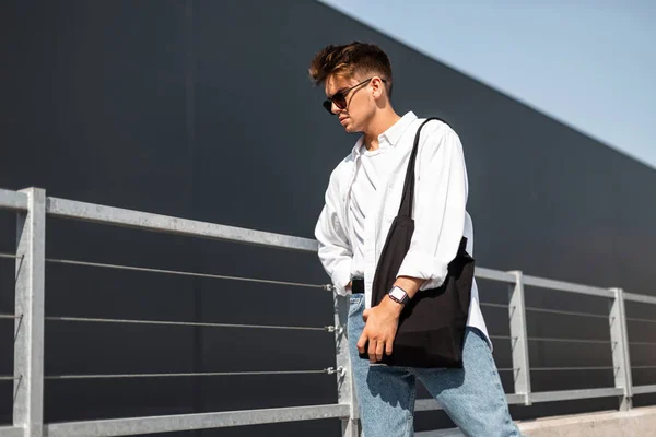 Stijlvolle jonge man in Vintage wit overhemd in modieuze zonnebril trendy blauwe jeans met een doek zwarte tas poseren op een zonnige dag in de buurt van een grijs gebouw buiten. Moderne kerel model. Jeugd zomer stijl. — Stockfoto