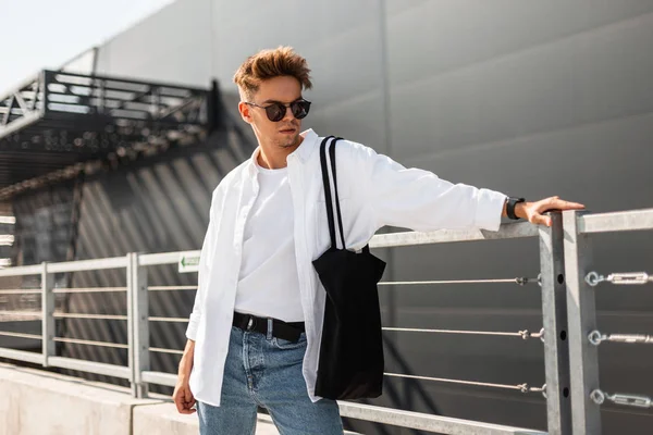 Mooie moderne jonge man model in een elegant wit overhemd in jeans in een t-shirt in zonnebrillen met een tas staat in de buurt van een metalen reling in de stad op een zonnige dag. Amerikaanse hipster Guy. Zomer mode. — Stockfoto