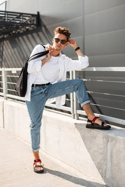 Homem hipster jovem na moda em roupas elegantes brancas e jeans em sandálias de couro vermelho em óculos de sol com um saco de tecido descansam perto de um edifício cinza em um dia ensolarado. Um americano. Rua juventude moda . — Fotografia de Stock