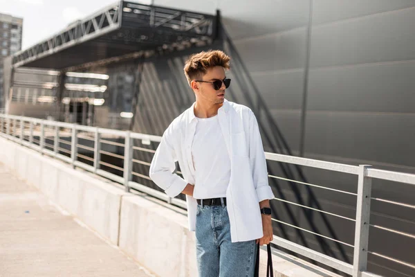 Stijlvolle knappe jonge man hipster in zonnebrillen in modieuze kleding met trendy kapsel met tas poseren in de buurt van een modern gebouw op een zonnige dag buiten. Aantrekkelijk Urban Guy model. Zomer stijl. — Stockfoto