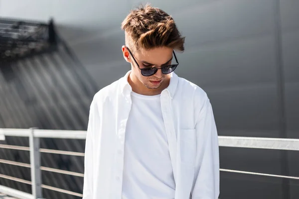 Knappe moderne jonge man in modieuze witte kleding in een stijlvolle zwarte zonnebril met trendy kapsel staat op een heldere zonnige dag in de buurt van een grijs gebouw in de stad. Amerikaanse kerel model. Zomer stijl — Stockfoto