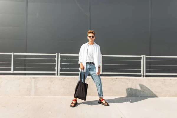 Mladý módní model moderního muže v bílé košili v modrých stylových džínách v slunečních brýlích s černým pytlem ve městě za jasného slunečného dne. Hezkej alternativců. Letní pohled. Mládežnická móda. — Stock fotografie