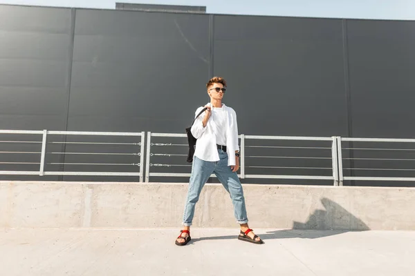 Moderne jonge man model in modieuze kleding in een zonnebril in stijlvolle rode lederen sandalen met een tas staat op een zonnige zomer dag in de buurt van een grijs gebouw. Urban hipster Guy. Trendy herenkleding. Streetstyle — Stockfoto