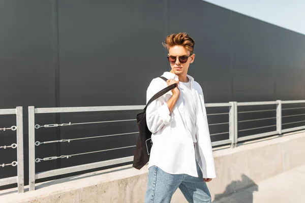 Cool Young Man model in een elegante witte shirt in blauwe jeans in stijlvolle zonnebril met zwarte doek zak is poseren in de buurt van een metalen reling in de stad op een zonnige dag. Amerikaanse hipster Guy. Zomer stijl. — Stockfoto