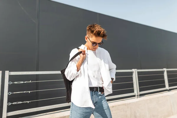 Attractive young man model in summer white fashionable clothes in sunglasses with a cloth black bag stands on a sunny day near a gray building in the city. Handsome guy hipster. European youth style.