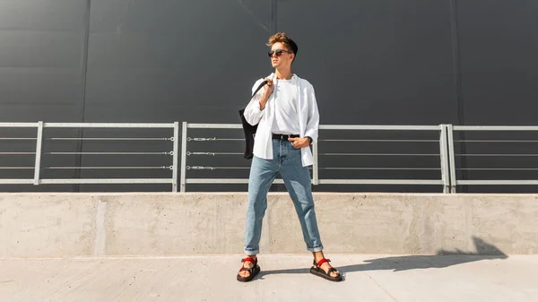 Jonge Amerikaanse man model in stijlvolle kleding in een zonnebril in rood lederen sandalen met een vintage zwarte tas staat op een heldere zonnige dag in de buurt van een grijs gebouw. Urban hipster Guy. Modieuze herenkleding. — Stockfoto