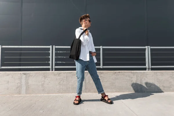 Jonge Europese man hipster in een wit overhemd in blauwe stijlvolle jeans in een donkere zonnebril met een zwarte tas staat op een zonnige dag in de stad. Moderne Urban Guy model. Modieuze zomerlook. Jeugd mode. — Stockfoto