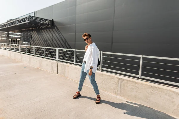 Молодой хипстер в белой модной рубашке в синих стильных джинсах в летних винтажных сандалиях в солнечных очках стоит в солнечный летний день рядом с современным зданием на улице. Модель модного парня . — стоковое фото