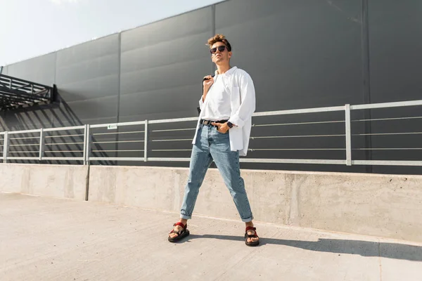 Nova coleção de roupa masculina de verão na moda. Homem hipster jovem atraente em roupas elegantes em óculos de sol vintage com um penteado com uma bolsa em sandálias posa na cidade em um dia ensolarado de verão . — Fotografia de Stock