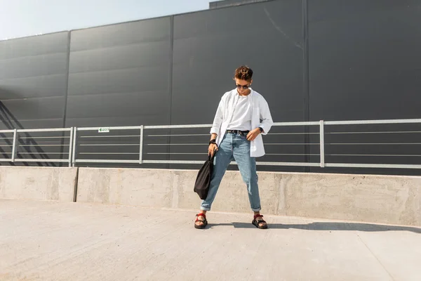 Joven modelo de hombre americano en una camisa blanca en vaqueros azules con estilo en gafas de sol con una bolsa negra pasea por la ciudad en un día soleado. Un hipster atractivo. Elegante look veraniego. Moda juvenil . — Foto de Stock