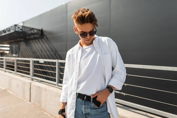 Trendy Young Man hipster in stijlvolle kleren in zwarte zonnebrillen met een kapsel rust op de straat op een zonnige dag. Aantrekkelijk Guy model buitenshuis. Nieuwe collectie zomer herenkleding. Modieuze look. — Stockfoto
