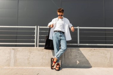 Bir çanta ile şık kırmızı deri sandalet içinde güneş gözlüğü şık yaz beyaz giysiler içinde modern genç adam hipster gri bir bina yakın güneşli bir günde duruyor. İyi adam. Trendy erkek giyim. Sokak stili