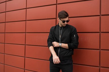 Trendy saç tarzı ile şık güneş gözlüğü moda ceket Amerikan güzel genç hipster adam bir yaz gününde şehirde yürür. Açık havada kırmızı binanın yakınında modern adam modeli. Sokak stili.