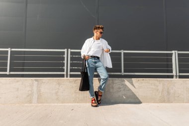 Bir kumaş çanta ile koyu güneş gözlüğü kırmızı Sandalet mavi kot klasik bir gömlek güzel genç adam hipster gri bir binanın yakınında sokakta dinleniyor. Avrupa adam modeli. Yaz trendy erkek giyim.