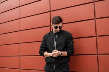 Şık güneş gözlüğü siyah ceket genç şık hipster adam gümüş saat düzeltir. Kırmızı bir binanın yakınındaki şehirde şık yakışıklı model adam açık havada. Trendy erkek giyim.