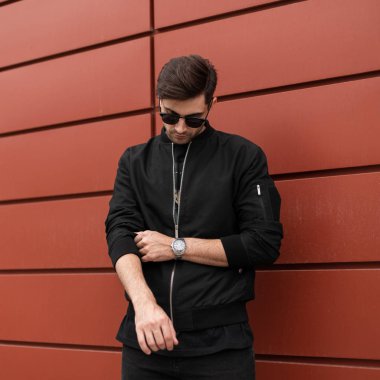 Trendy saç tarzı ile şık güneş gözlüğü moda ceket yakışıklı genç hipster adam bir yaz gününde şehirde yürür. Amerikan adam model kırmızı bina açık havada yakın poz. Sokak stili