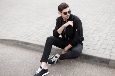 Trendy güneş ayakkabıları siyah kot şık bir ceket yakışıklı genç hipster adam yaz gününde bir sokakta kaldırım üzerinde oturuyor. Çekici adam Model açık havada rahatlatıcı.