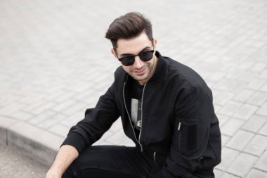Kot siyah moda ceket şık güneş gözlüğü mutlu genç hipster adam güneşli bir yaz gününde bir sokakta bir karo üzerinde oturan dinleniyor. Joyful Amerikan Model adam rahatlatır açık havada.