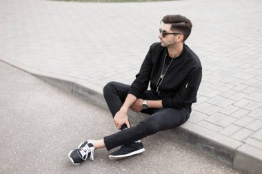 Trendy güneş ayakkabıları siyah kot şık bir ceket içinde kentsel genç hipster adam yaz gününde bir sokakta kaldırım üzerinde oturuyor. Yakışıklı adam Model açık havada rahatlatıcı.