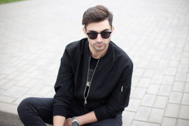 Karanlık güneş gözlüğü şık siyah giysiler içinde genç model adam dinlenmek parlak bir yaz gününde şehirde bir çini oturuyor. Yakışıklı Amerikan adam modeli hafta sonu açık havada sahiptir. Trendy erkek giyim.
