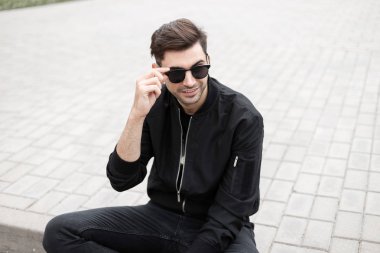 Kot şık bir siyah ceket içinde trendy güneş gözlüğü gülümseme ile pozitif genç hipster adam parlak bir yaz gününde şehirde kaldırımda oturuyor. Neşeli adam düzleştirir güneş gözlüğü.