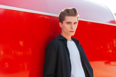 Klasik bir siyah gömlek şık bir T-shirt bir moda saç modeli ile genç zarif adam parlak bir yaz güneşli bir gün kırmızı metal duvar yakınında duruyor. Ciddi adam açık havada dinleniyor. 