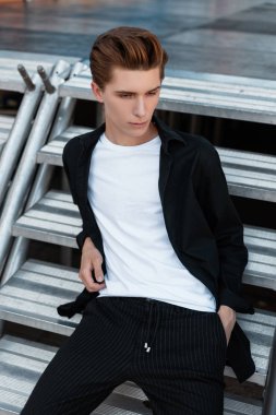 Şık bir moda saç stilinde şık genç adam modern bir gümüş metal merdiven şehirde oturan rahatlatır. Yakışıklı kentsel adam bir yaz gününde rahatlatıcı sahiptir.