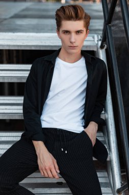 Modern çekici genç adam bir T-shirt içinde şık bir siyah gömlek trendy pantolonu içinde bir metal merdiven üzerinde oturan oturma içinde zarif bir saç modeli ile şehir. Yakışıklı adam dinlenmek üzerinde bu sokak.