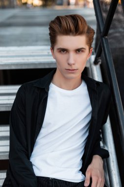 Beyaz bir tişört zarif bir siyah gömlek şık bir saç modeli ile genç yakışıklı adam şehirde bir metal merdiven oturuyor. Çekici adam. Şık erkek giyim.
