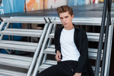 Zarif trendy giysi şık bir saç modeli ile modern genç adam sokakta modern bir gümüş metal merdiven oturan rahatlatır. Çekici adam bir yaz gününde rahatlatıcı sahiptir. Erkek giyim.