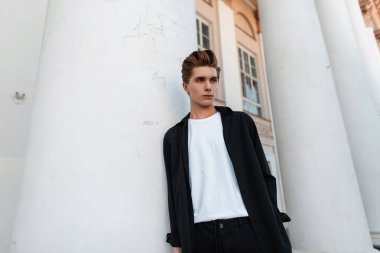 Trendy bir saç modeli ile çizgili pantolon beyaz şık t-shirt bir moda siyah gömlek çekici genç adam beyaz bir Vintage sütun yakın dayanır. Bir yaz gününde şehirde yakışıklı adam.