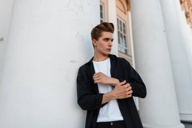 Şık bir saç stili ile bir t-shirt trendy siyah gömlek Avrupa yakışıklı genç adam şehirde sütunlar ile beyaz bir Vintage bina yakın pozlar. Zarif adam moda modeli. Şık erkek giyim.