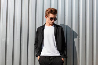 Zarif trendy giysi şık bir güneş gözlüğü bir saç modeli ile modern genç adam hipster sokakta Vintage metal duvar yakın rahatlatır. Çekici adam hoşlanır rahatlatıcı üzerinde bir güneşli yaz gün. 