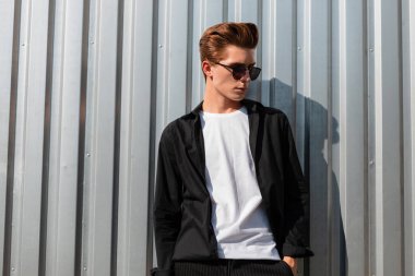 Moda güneş gözlüğü zarif giysiler içinde Trendy saç modeli ile Avrupa şık genç adam güneşli bir günde şehirde parlak bir metal duvar yakın pozlar. Yakışıklı hipster adam güneş sahiptir. Erkek giyim.