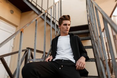 Zarif pantolon bir T-shirt şık bir siyah gömlek moda saç modeli ile çekici genç adam eski bir demir merdiven oturuyor rahatlatır. Şehir açık havada yakışıklı adam. Trendy erkek giyim.